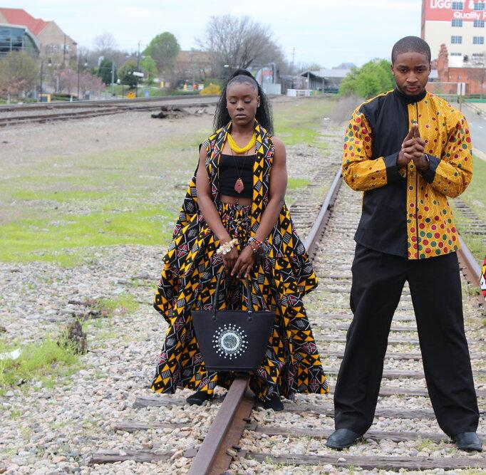Two individuals standing on train tracks wearing yellow and black traditional West African attire.