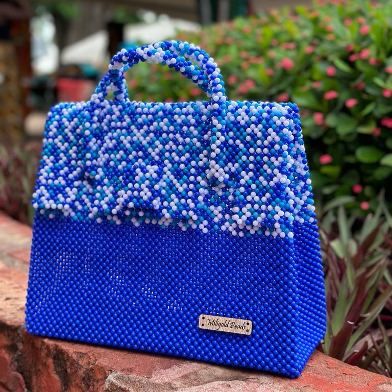 A blue and white Miligold handbag made of plastic beads, displayed on a brick surface with plants in the background.